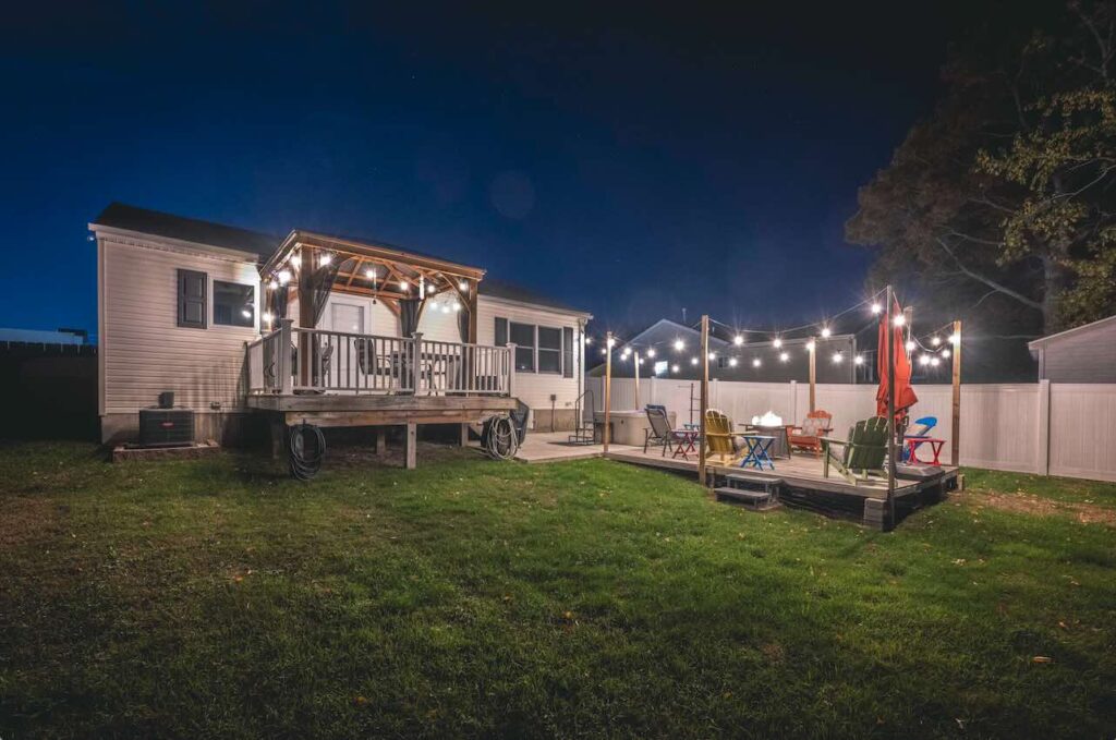 A cozy Cape May, New Jersey backyard at night with a lit pergola deck, string lights, colorful Adirondack chairs, and a fire pit—creating a warm, inviting coastal atmosphere for relaxing evenings.