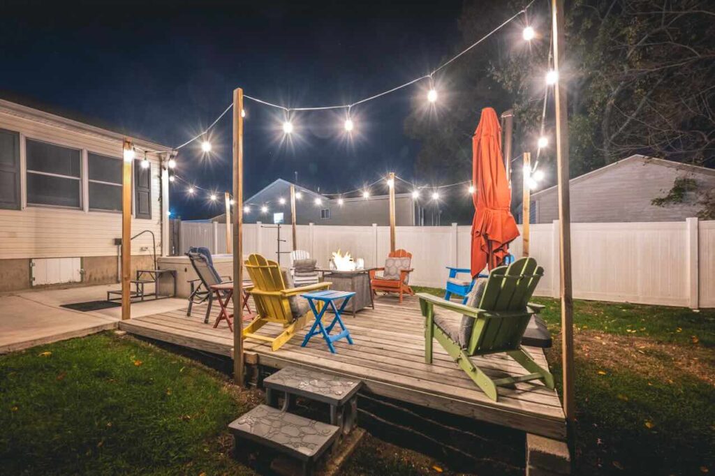 A charming Cape May, New Jersey backyard patio at night with colorful Adirondack chairs around a fire pit, glowing string lights overhead, and a cozy, beach-town ambiance perfect for relaxing evenings.