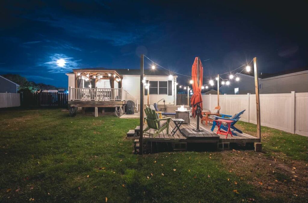 A peaceful Cape May, New Jersey backyard at night featuring a lit pergola deck, string lights over colorful Adirondack chairs and a fire pit, with the full moon glowing in the sky above the cozy coastal home.