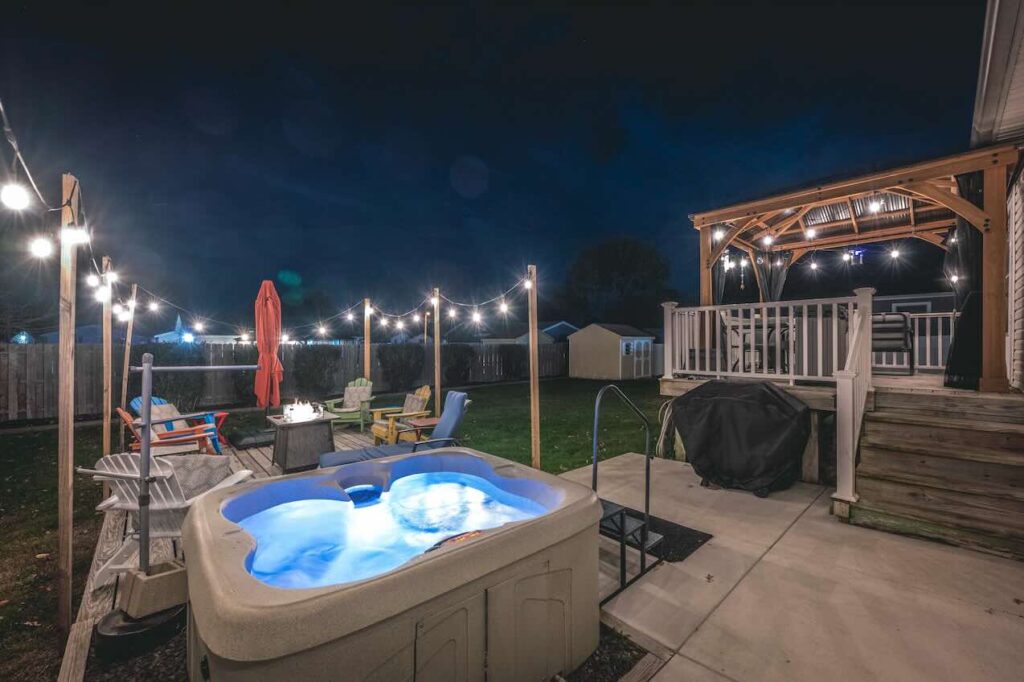 A cozy Cape May, New Jersey backyard at night featuring a bubbling hot tub, string lights, colorful Adirondack chairs around a fire pit, and a covered patio with seating—creating a relaxing coastal retreat atmosphere.